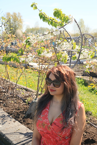 A Fijian model in a garden in 
Springtime on a sunny day. She is wearing long brown wavy hair, sunglasses, makeup, and a red pattern sleeveless dress.