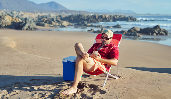 mature man sitting alone relaxed holding and looking a smartphone enjoying beach and nature