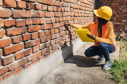 A professional female engineer in a yellow hard hat is examining a brick wall for structural integrity, holding a clipboard