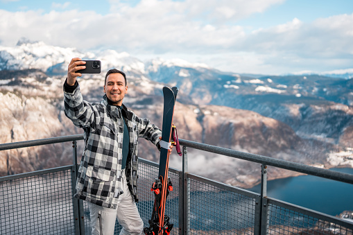 A mid adult Asian male skier captures a selfie against a scenic mountain landscape, dressed in casual winter attire with ski equipment.