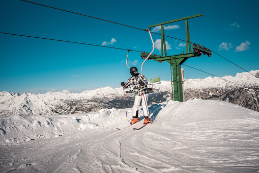 A mid adult Asian male skier getting off a ski lift on a snowy mountain slope. He is wearing a plaid jacket and orange pants, with ski equipment on his feet and a helmet for safety.