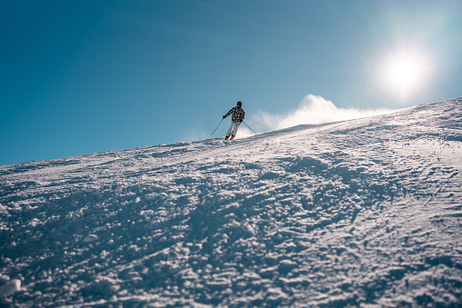 A mid adult Asian male skier in motion descending a snowy mountain slope under clear skies with sunlight glinting off the snow.