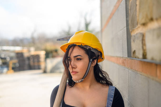 A woman wearing a yellow helmet and safety glasses is working on a construction site. She is wearing a blue apron and is bending over to pick up a brick