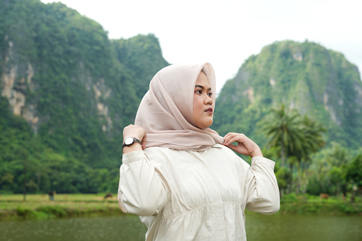 A female model wearing a hijab is posing in nature with a village and green mountains in the background