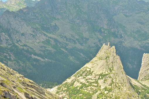 A high steep rock with a pointed peak surrounded by high mountains on a sunny summer day. The thin brother of the Parabola rock, Ergaki Natural Park, Krasnoyarsk Territory, Siberia, Russia.