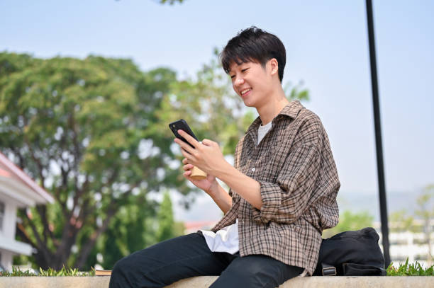 istock A cheerful young Asian male college student sits on a stone bench in a park, using his smartphone. 2149759096