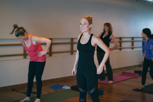 A smiling young, athletic woman stands on her mat, participating in a community yoga class in a fitness studio.