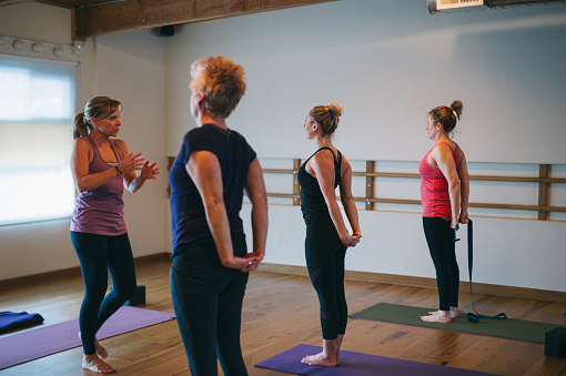 An instructor leads a multi-ethnic group of ladies in a shoulder stretch during a yoga class in a fitness studio.