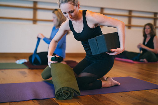 An athletic woman of Caucasian descent kneels on the floor and rolls up her yoga towel and mat after exercising in a community fitness studio.