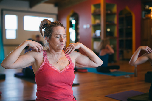 An athletic Caucasian woman sits on a mat with her hands on her shoulders, doing breathing exercises during a community yoga class in a fitness studio.
