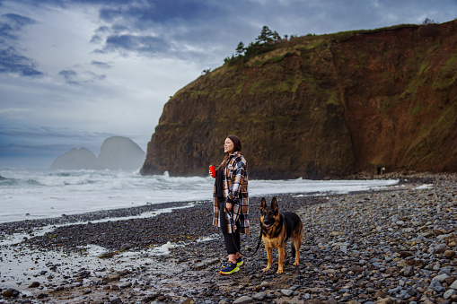 A woman dressed in warm clothing enjoys a rainy Winter day at the Oregon Coast with her companion, a well trained German Shepard dog.