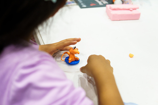 A girl is making a toy out of clay. She is wearing a purple shirt. The toy is orange and blue
