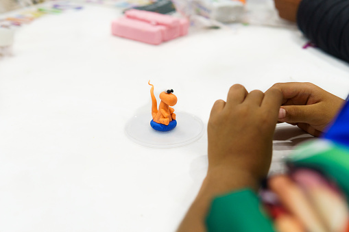 A child is making a dinosaur out of clay. The dinosaur is orange and blue and is sitting on a white surface