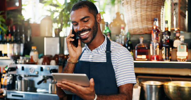istock Tablet, bartender and black man on cellphone call, conversation and order alcohol stock, store delivery or inventory. Restaurant checklist, phone and business owner consulting on drinks supply chain 2149772125