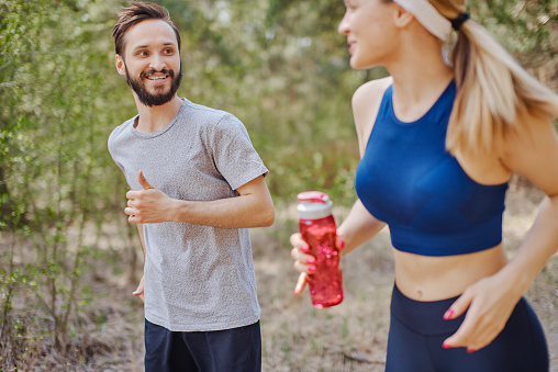 Close up portrait of two people jogging in the forest in spring, exercising in the park. Woman is holding a water bottle in hands, man is smiling at her