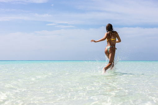 Back view of carefree woman splashing water while running through sea in summer day. Copy space.