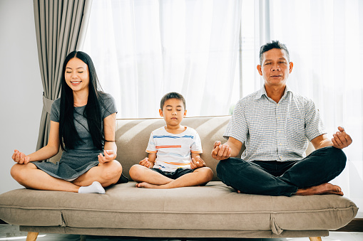 Calmness and family bonding as parents and their young son practice yoga on a sofa. Illustrating the serenity of meditation fostering harmony and peace within the household.