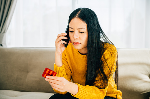 A young Asian woman unwell takes medicine consults a doctor via phone on a couch at home. Illustrating medical consultation medication and social distancing amid coronavirus.