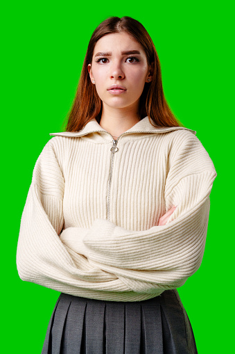 A young woman stands with a confident posture, arms folded across her chest, wearing a cream-colored zip-up sweater and a dark pleated skirt. Her expression is serious and contemplative, reflecting a sense of determination or thoughtfulness.