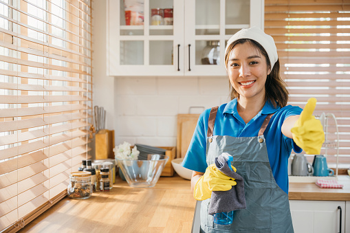 Smiling woman in uniform apron and rubber glove ready for home cleaning. Emphasizing housekeeping and hygiene with spray bottle. Clean disinfect home care. maid with liquid.