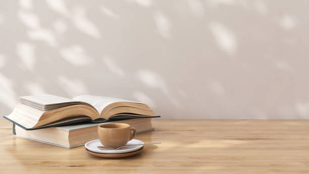 Brown wooden countertop table with opened book and coffee cup in dappled sunlight, leaf shadow on white wall in background