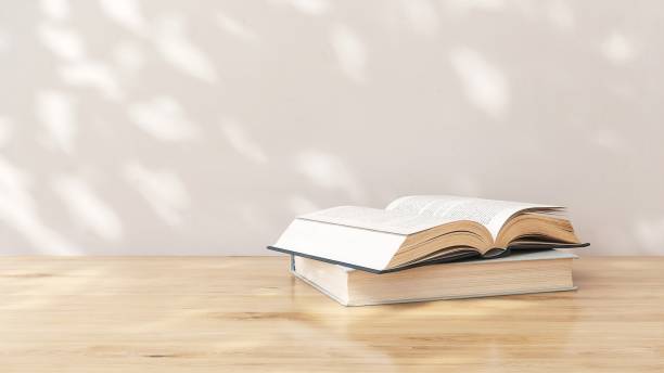 istock Brown wooden countertop table with opened book in dappled sunlight, leaf shadow on white wall in background 2149775483