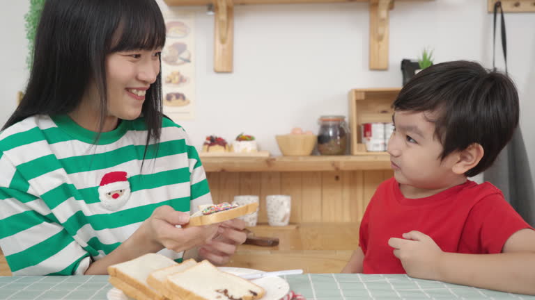 Young boy making an chocolate sandwiches at the breakfast table with his Asian mother in the kitchen