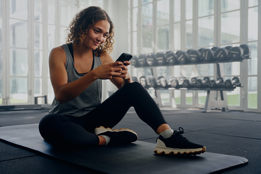 Young multiracial woman wearing sportswear sitting and using mobile phone while taking a break at the gym