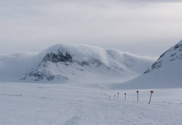 A nordic ski tour trail in the Sylarna mountains in Sweden. Red cross trail markers line the trail and clouds roll over the mountains in the background.