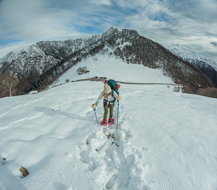 A female adventurer in red snow shoes is snowshoeing up a steep snowy hill, showing clear determination. She's focused on moving through the untouched snow, with mountains and a cloudy sky in the distance. Dressed in winter gear, carrying a backpack, and using trekking poles, she's equipped for a tough journey.