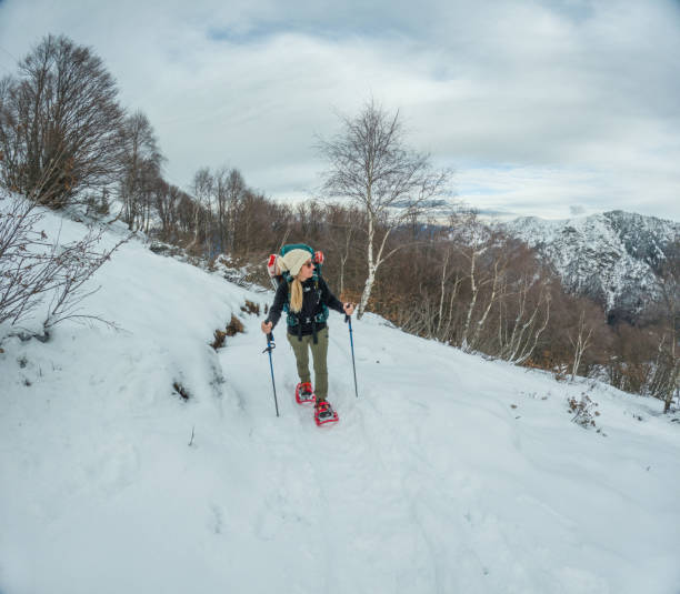 A female adventurer in red snow shoes is snowshoeing up a steep snowy hill, showing clear determination. She's focused on moving through the untouched snow, with mountains, trees and a cloudy sky in the distance. Dressed in winter gear, carrying a backpack, and using trekking poles, she's equipped for a tough journey.
