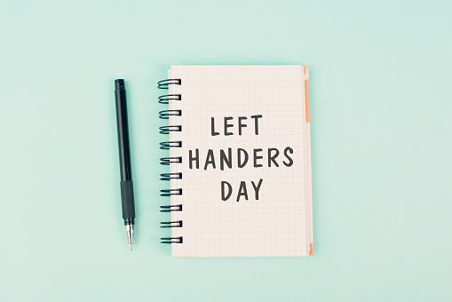 Left handers day is standing on a notebook, writing with the left hand, pen and table