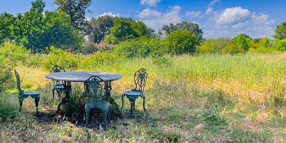 A beautiful garden with an elegant Ottoman-style marble table surrounded by metal chairs. Spring blossoms with lush meadows and trees under a blue sky with white clouds. Serene background with copy space.