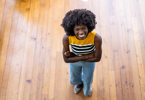 A cheerful Afro-Caribbean woman standing on a wooden floor with her arms crossed is looking up to smile at the camera