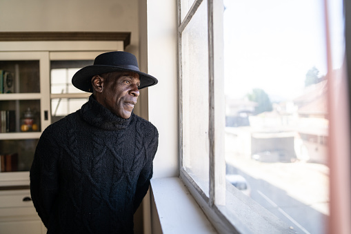 A senior Afro-Caribbean man is standing beside a large window, looking outside to the bright sunny view