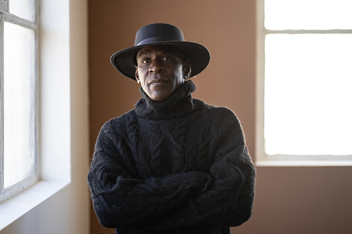 An African-American man wearing a heavy sweater and a cowboy hat is standing with his arms crossed and looking seriously at the camera