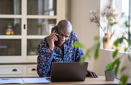 An Afro-Caribbean man wearing a smart shirt is sitting at his desk, talking on a mobile phone while looking at his laptop screen