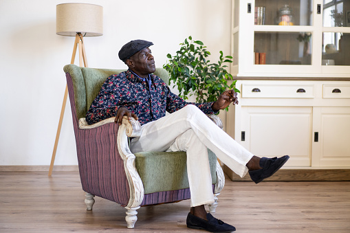 A full length shot of a mature African-American man wearing a smart snazzy shirt, white trousers and a flat cap while relaxing in an armchair in a modern room