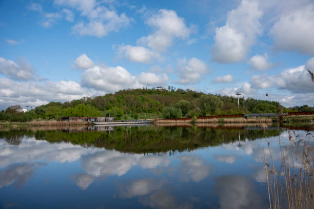 A pond in which snow-white clouds are reflected, a hill covered with forest and a metal bridge. Landscape of a city park.