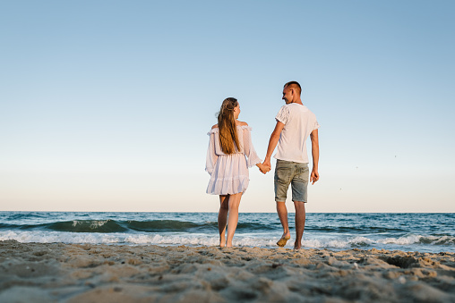 Female and male walk barefoot on beach ocean and enjoy sunny summer day on vacation. Man and woman walking on sand sea. Happy couple in love holding hand go on seashore. Spend time together. Back view