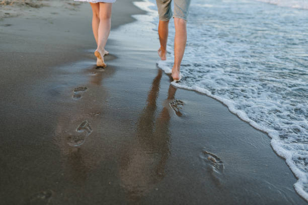 Down view of bare feet on sand beach. Couple in love walking barefoot on water with big waves on seashore. Man and woman walk on sea. Spending time together. Bottom view on legs. Back view. vista de abajo de los pies descalzos en la playa de arena. pareja de enamorados caminando descalzos sobre el agua con grandes olas en la orilla del mar. el hombre y la mujer caminan sobre el mar. pasar tiempo juntos. vista inferior de las piernas. vista po