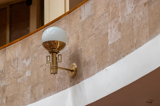 A wall-mounted lamp with a white dome on a gold fixture adorns a marble wall.