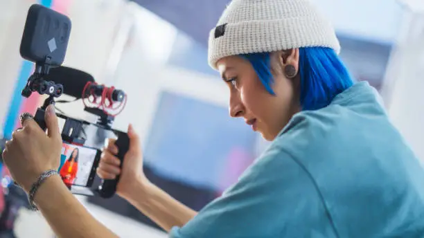 Focused Young Caucasian Female Cinematographer With Blue Hair Adjusts Camera Settings On Film Set, Capturing Vibrant, Creative Video Content In A Professional Studio Environment.