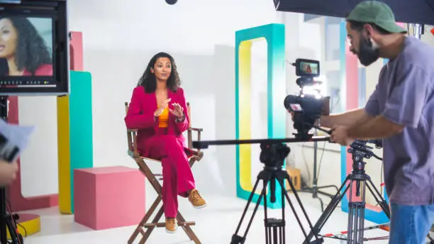 Vibrant Film Set Captures Young South Asian Female Host In A Pink Suit Engaging In A Lively Discussion, Surrounded By Colorful Set Design And Busy Crew Handling Video Equipment For A Dynamic Shoot.