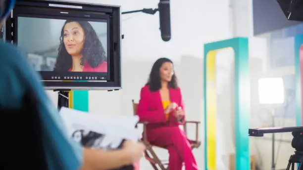 Vibrant On-set Image Of A Young Black Female Host In A Pink Suit, Engaging In A Lively Discussion During A Tv Show Recording, Surrounded By Modern Videography Equipment And Colorful Set Design.