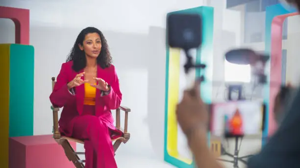 Vibrant Scene Captures A Black Female Host In A Pink Suit, Engaging Animatedly In A Colorful Studio Setup, Surrounded By Modern Geometric Props, During A Lively Video Recording Session For A Talk Show