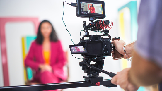 Vibrant Scene Captures Young Asian Female Host In Pink Blazer On Film Set, Engaging In Lively Discussion, With Focus On Professional Video Equipment And Dynamic Set Design.