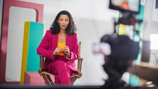 Young Black Female Host In Vibrant Pink Suit Engaged In Lively Discussion On Colorful Set, Captured On Camera For A Modern Talk Show, Emphasizing Dynamic Storytelling And Visual Appeal.