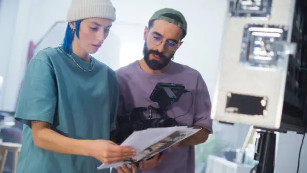 Young Caucasian Female And Middle Eastern Male Cinematographers Review A Script By A Camera On A Busy Film Set, Focusing On Video Production For A Documentary Or Tv Show.