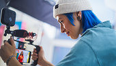 Young Caucasian Female Cinematographer With Blue Hair Adjusts Camera Settings On Film Set, Capturing Vibrant, Dynamic Footage For A Modern Advertising Campaign Or Documentary.